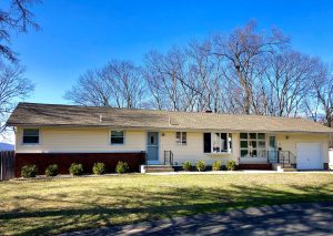 Color photo of a long ranch house with yellow siding, red brick below on the left, two entrances with short stairs and a garage.