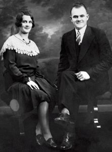 Historical black and white photo of a couple seated side by side, smiling and dressed formally.