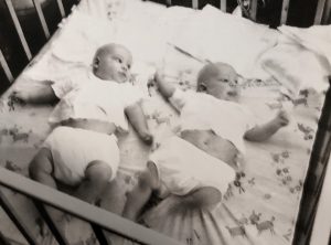Black and white photos of two babies, dressed identically, awake in a crib.